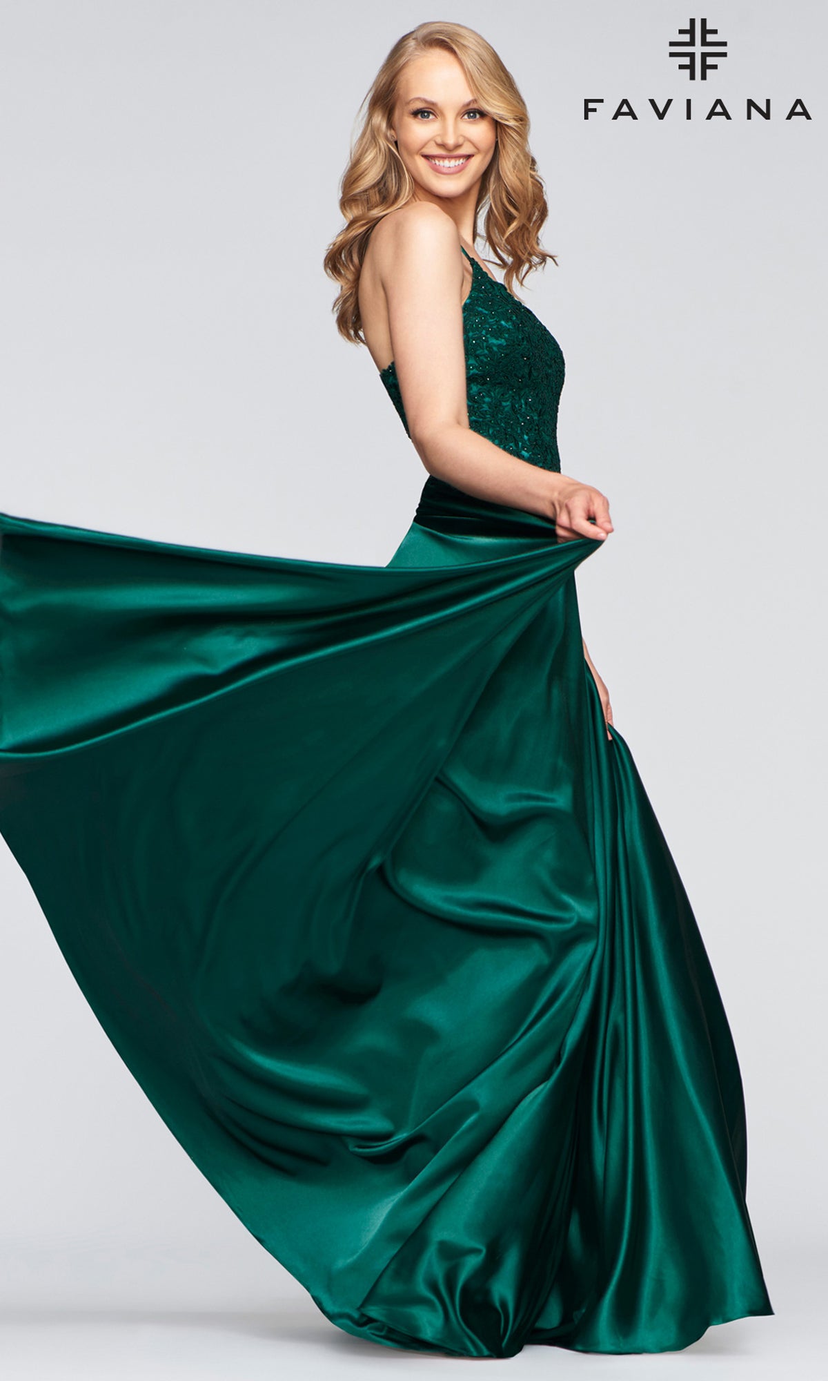 A woman smiles and poses, holding out the skirt of a green Faviana Lace-Bodice Long A-Line Prom Dress. The Faviana logo appears in the top right corner.